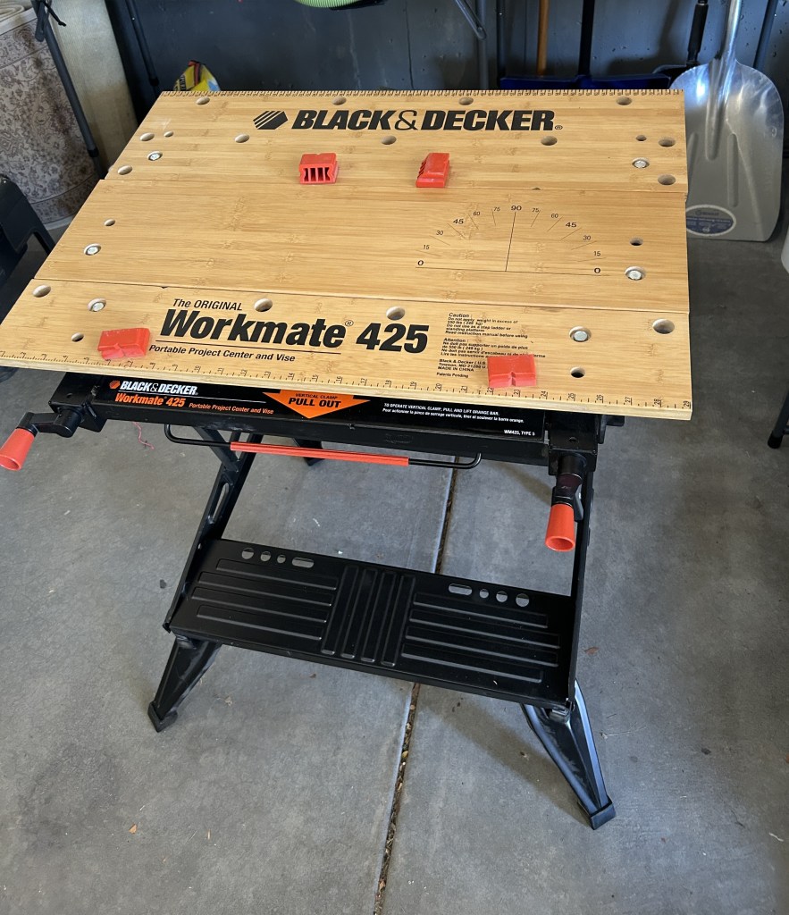 A Black & Decker Workmate 425 portable project center and vise, featuring a wooden top with measuring markings and red clamps, positioned on a garage floor.